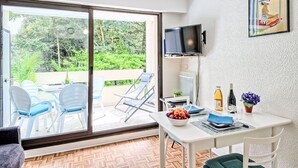 Une pièce dotée d’une porte-fenêtre coulissante donnant sur un jardin, une table blanche sur laquelle se trouve un saladier rempli de fruits, une bouteille de vin et un téléviseur fixé au mur.