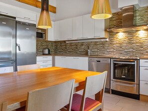 Cozinha moderna com eletrodomésticos de aço inoxidável, mesa de jantar de madeira e iluminação pendente.