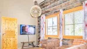 A cozy living room with a wooden door, a television mounted on the wall, a stone wall, and a large window with curtains.