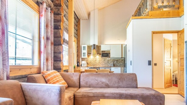 A modern log cabin interior with a large L-shaped sectional sofa, a wooden coffee table, and a kitchen area with stone backsplash and modern appliances.