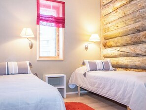 A room with two beds, a window with blinds, a nightstand, and a log wall.