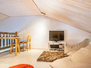 A cozy attic room with a wooden ceiling, a TV on a white stand, a sofa, a chair, and a small rug.