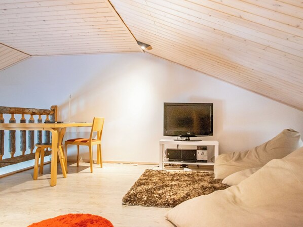 Una habitación cálida en el ático con techo de madera, un televisor sobre un soporte blanco, un sofá, una silla y una pequeña alfombra.