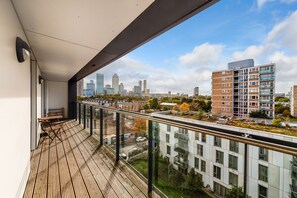 Ein Balkon mit Holzboden und Glasgeländer mit Blick auf eine Skyline moderner Gebäude und einen klaren Himmel.