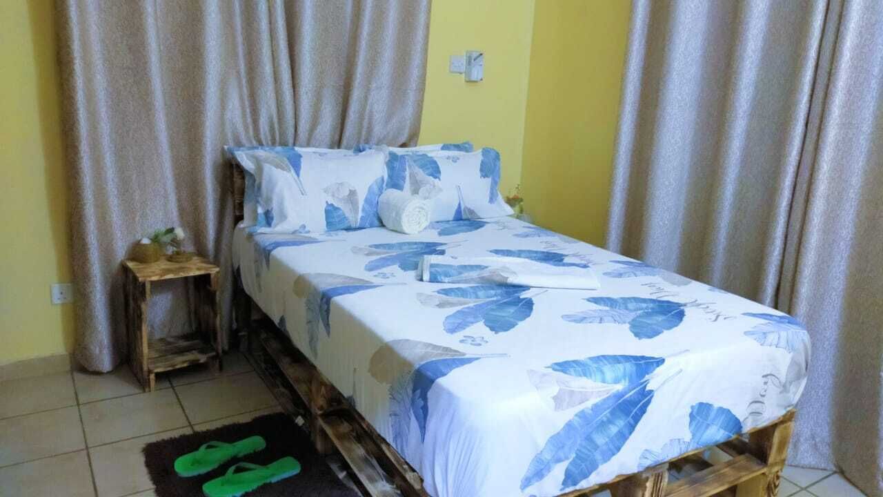 A single bed with a floral bedspread, a wooden nightstand with a lamp, and a pair of green slippers on a rug.
