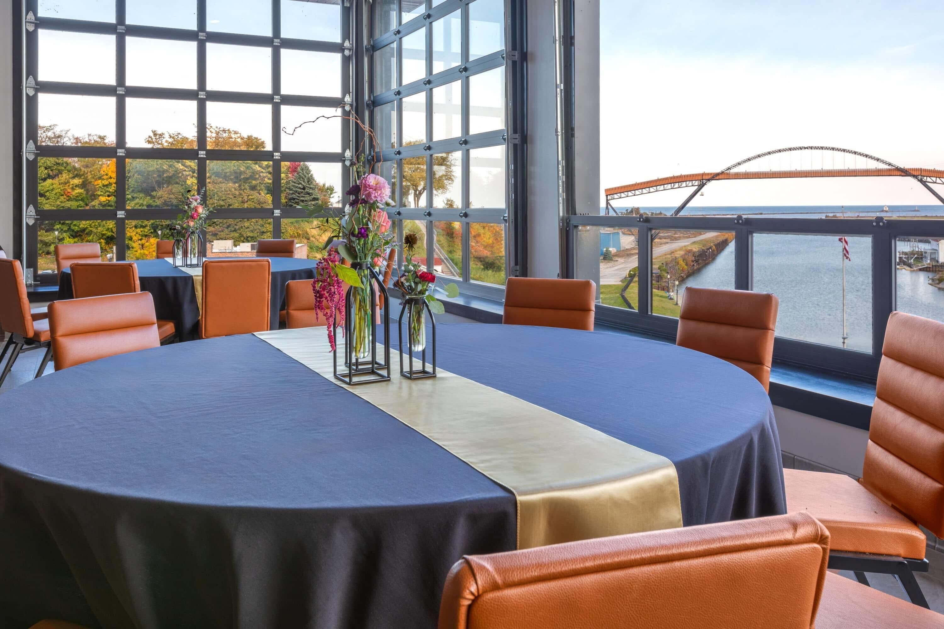 Una mesa redonda con mantel azul, rodeada de sillas naranjas, en una habitación con ventanas amplias que dan a un puente y al agua.