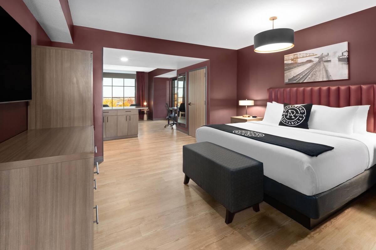A modern hotel room with a large bed, a TV, a desk, and a view of the cityscape.