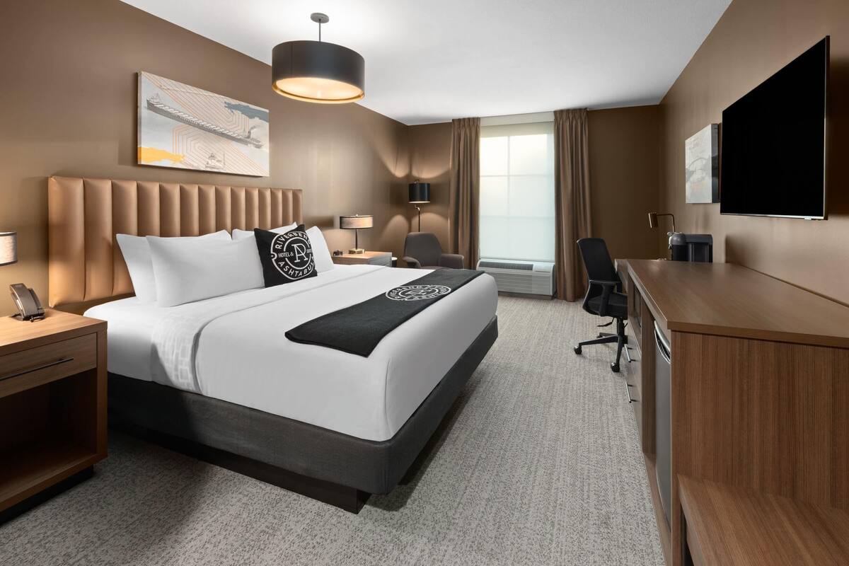 A hotel room with a large bed, a desk, a chair, a TV, and a window with curtains.