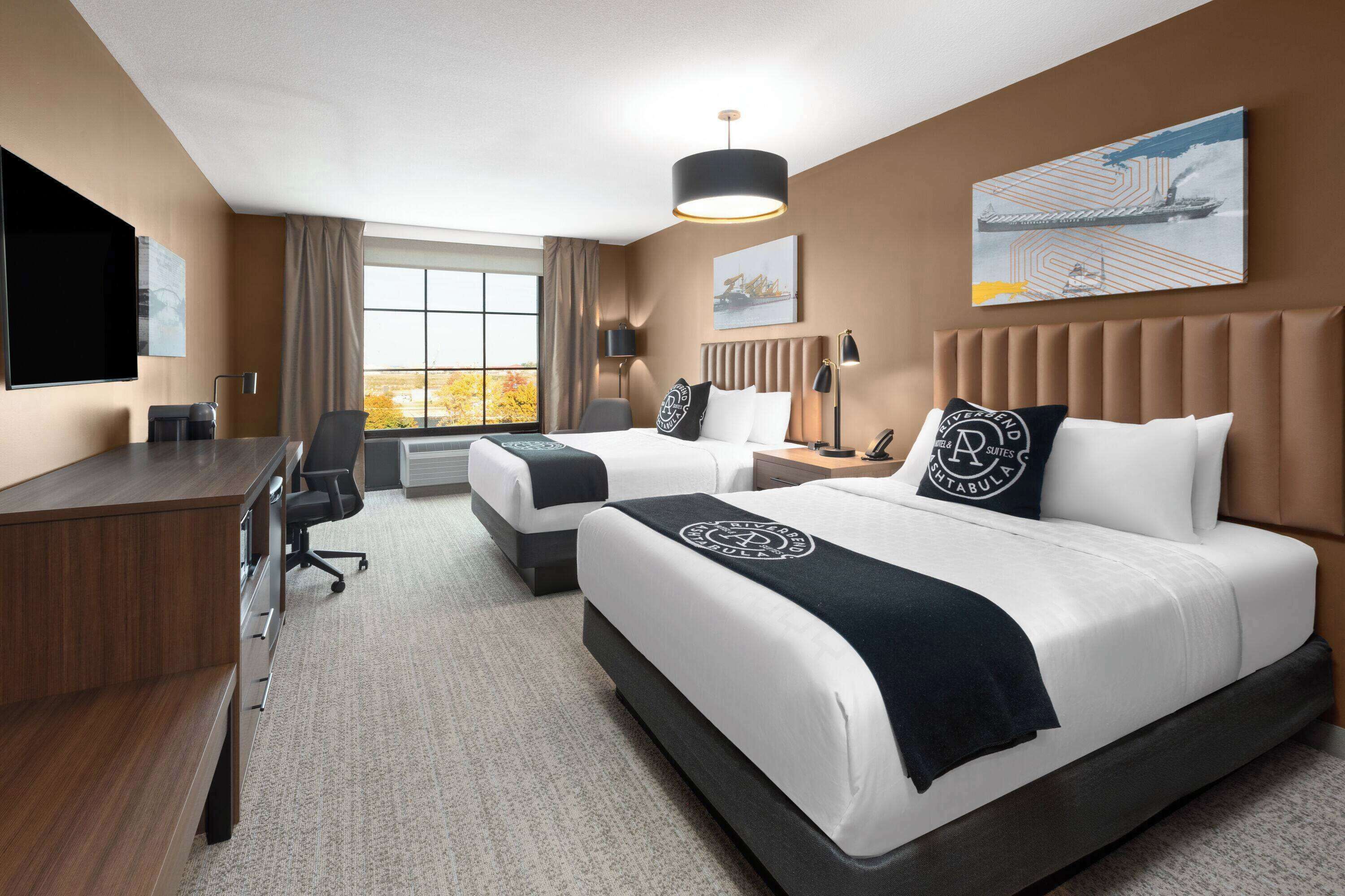 A hotel room with two beds, a desk, a chair, a TV, and a large window.