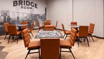 Un comedor con sillas y mesas naranjas, un mural de un puente, y un letrero que dice 'THE BRIDGE ROOM'.