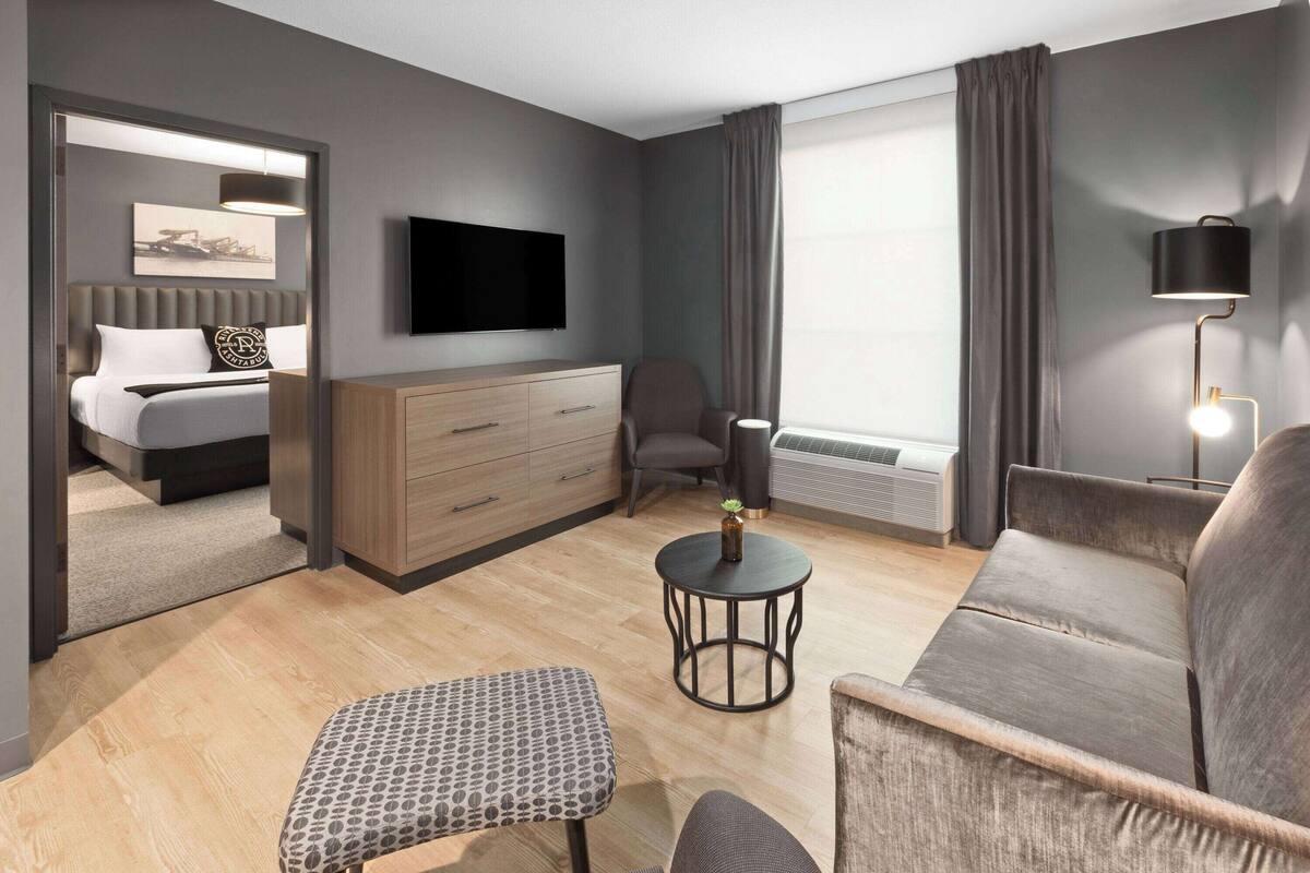 A modern hotel room with a bed, a TV, a desk, a chair, and a sofa.