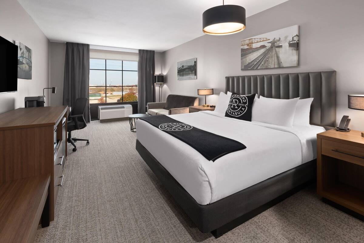 A modern hotel room with a large bed, a desk, a chair, a lamp, and a view of the cityscape.