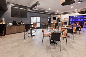 Un comedor moderno con varias mesas y sillas, una zona de cocina con mostrador y electrodomésticos, y un amplio ventanal con persianas.