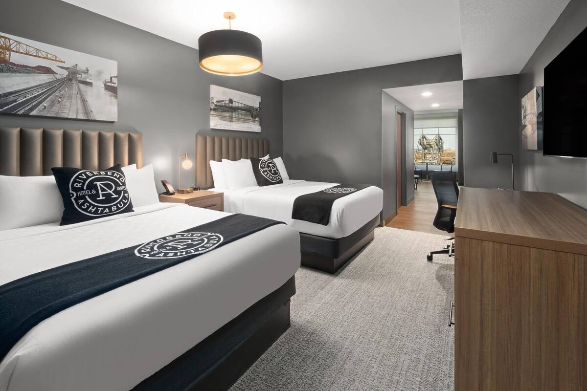 A modern hotel room with two beds, a desk, a TV, and artwork on the walls.