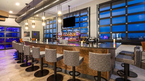 A bar area with a long counter, multiple stools, and a large window with a view. - Riverbend Hotel and Suites, Trademark Collection by Wyndham (Ashtabula)