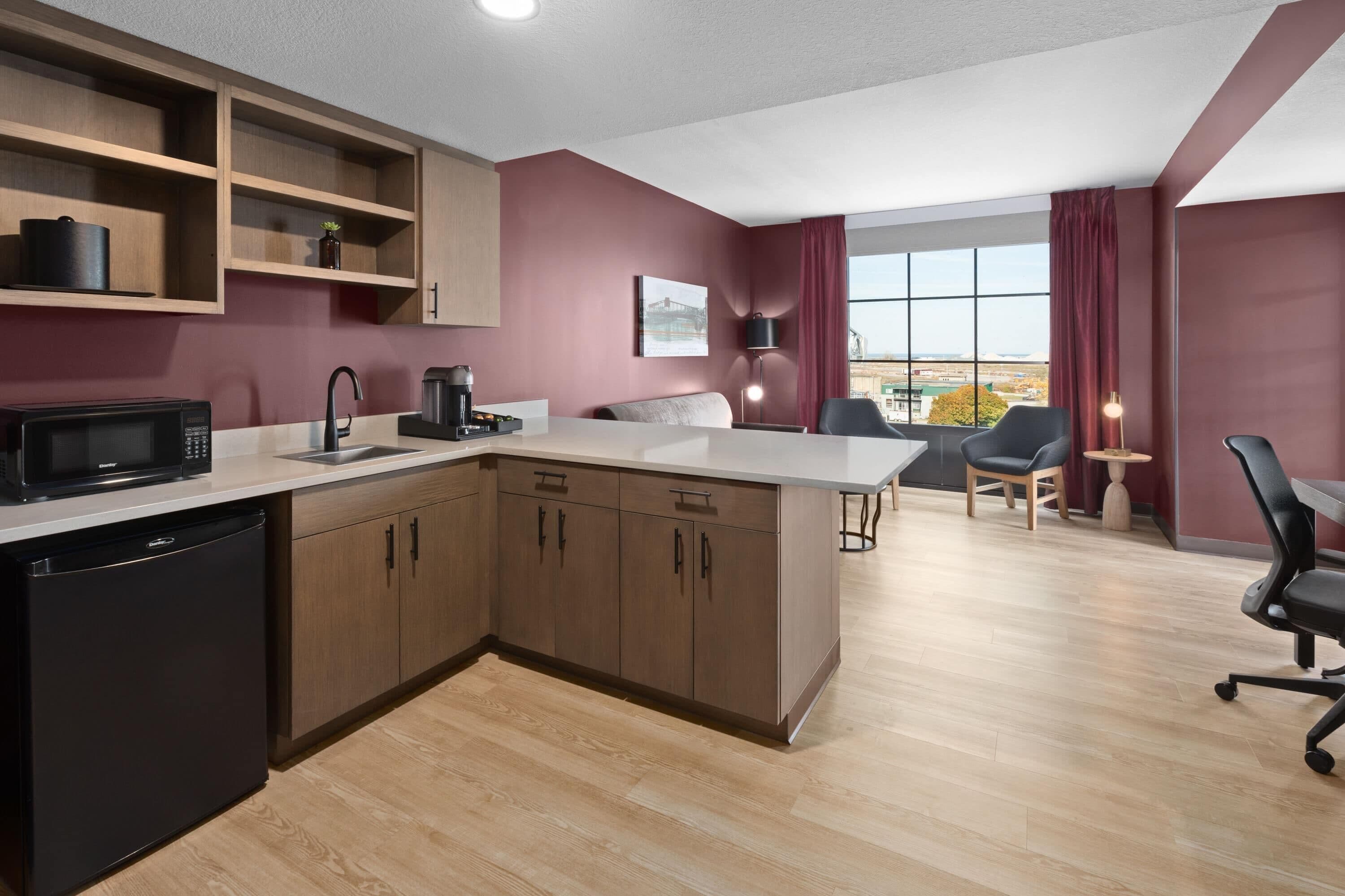 A modern kitchen with wooden cabinets, a black microwave, and a sink. There is a dining area with a table and chairs, and a large window offering a view.