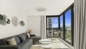 A modern living room with a grey sectional sofa, a small round table with two chairs, and a large window offering a view of buildings and greenery. - A-405 Stunning one bedroom apartment overlooking the pool (Cupecoy)