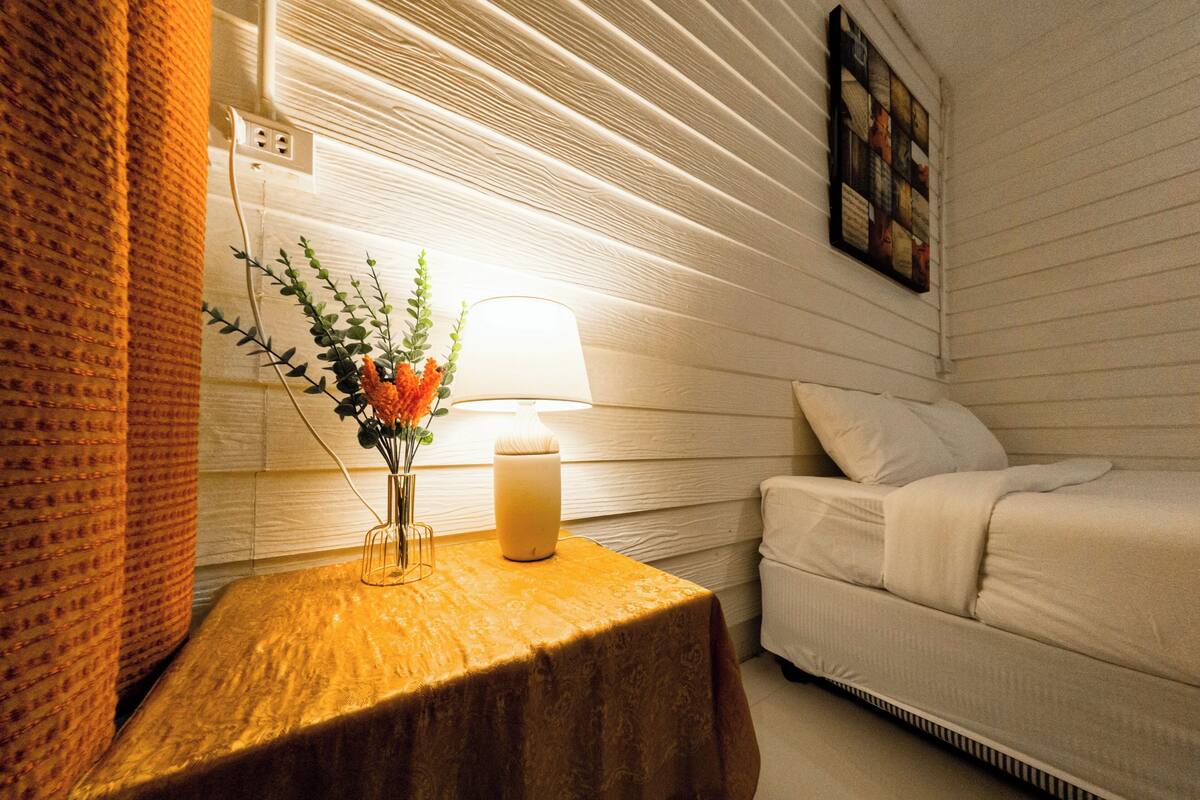 A room with a wooden nightstand, a lamp, a vase with flowers, a white bed, and a wall-mounted picture.