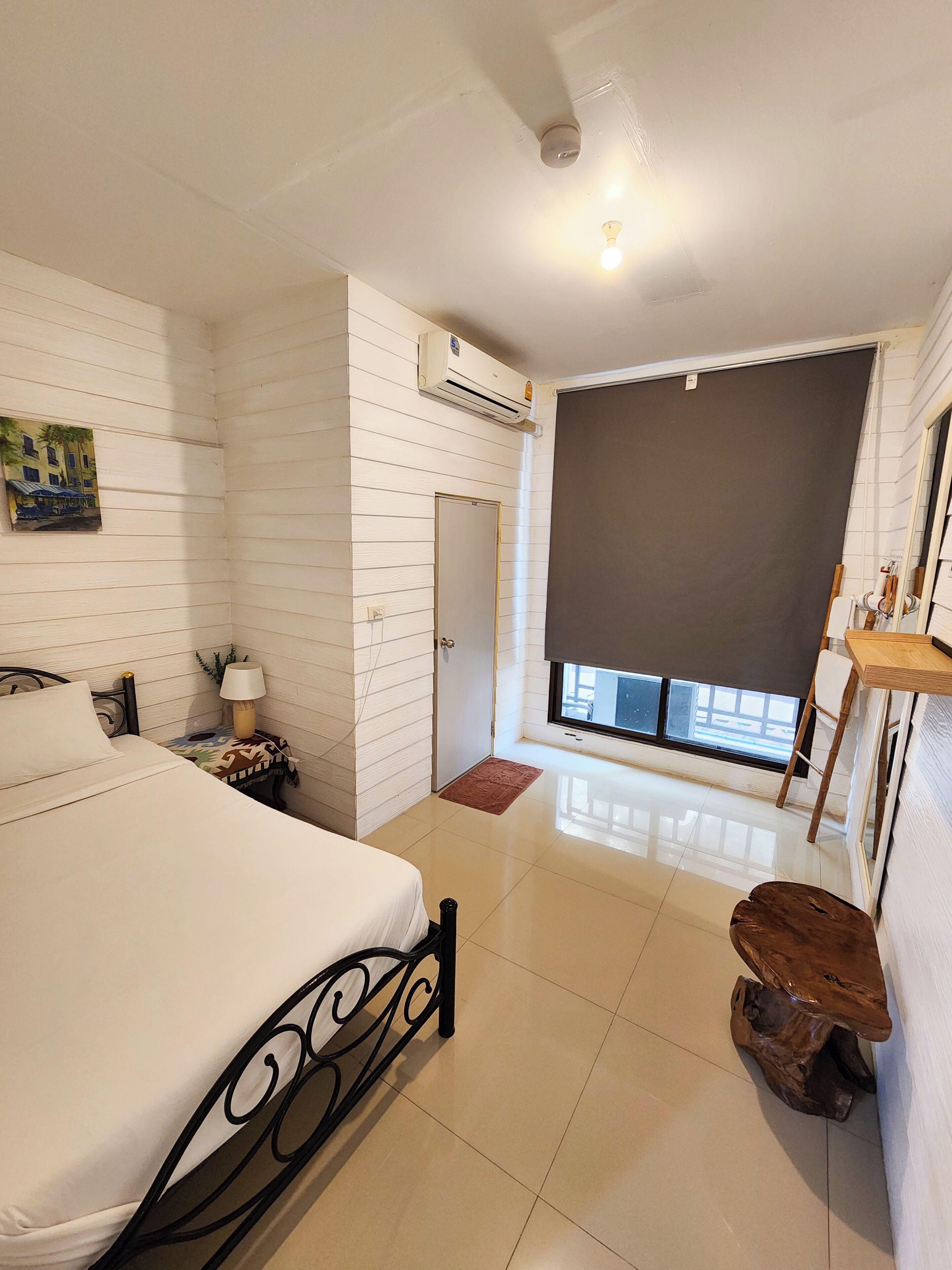 A bedroom with a bed, bedside table, lamp, air conditioning unit, and a window with a view.