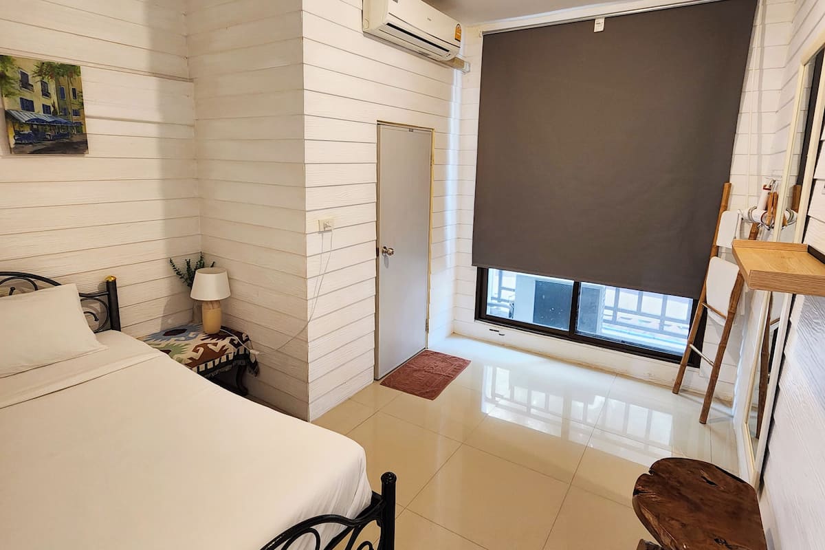 A bedroom with a bed, bedside table, lamp, air conditioning unit, and a window with a view.