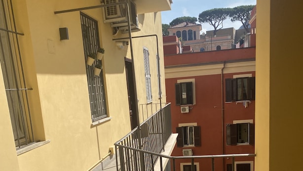 Un étroit couloir extérieur doté d’un sol carrelé, d’une rambarde métallique et offrant une vue sur les bâtiments adjacents.