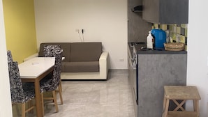 Une cuisine compacte avec un plan de travail gris, un four encastré et une machine à café. On y trouve un coin repas avec une table blanche et des chaises à motifs, ainsi qu’un canapé placé contre le mur.