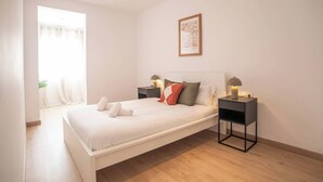 A neatly made bed with white linens and rolled towels, two bedside tables with lamps, a framed artwork on the wall, and a window with curtains. - 22CRC1039 Exquisite Apartment near the City Center (Barcelona)