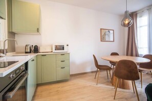 Une cuisine compacte équipée d’un four à micro-ondes, d’une bouilloire et d’une cuisinière, un coin repas avec une table et des chaises en bois, et un luminaire suspendu.