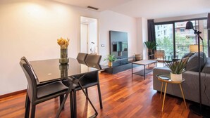 A modern living room with a dining area, a flat-screen TV mounted on the wall, and a large window providing a view of the city.