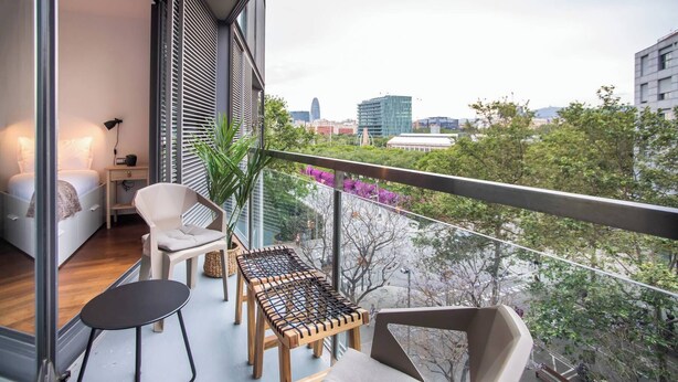 A balcony with a bed, a small table, and a chair, overlooking a cityscape with trees and buildings.