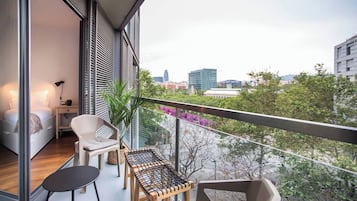 Een balkon met een bed, een tafeltje en een stoel, met uitzicht op een stadsgezicht met bomen en gebouwen.