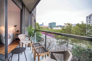 A balcony with a bed, a small table, and a chair, overlooking a cityscape with trees and buildings. - 42BADR1040 - New & Bright Apartment PobleNou (Barcelona)