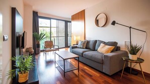 A modern living room with a grey sofa, a coffee table, and a TV mounted on the wall. There are potted plants and a lamp on a side table.