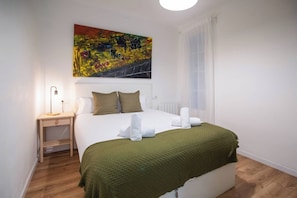 Habitación de hotel con cama, escritorio, lámpara, un cuadro en la pared y ventana con cortinas.