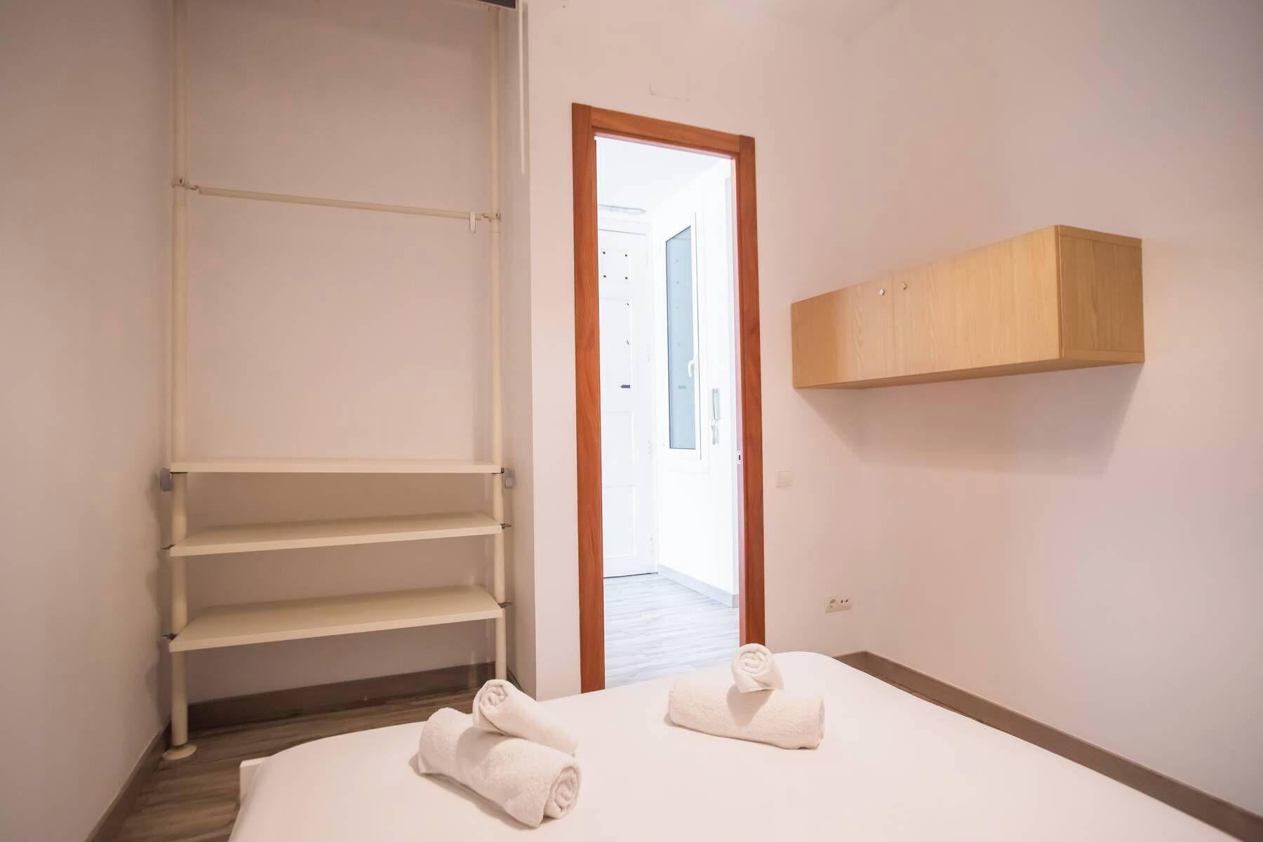 A hotel room with a bed, two rolled towels, a wooden wardrobe, and a white shelf unit.
