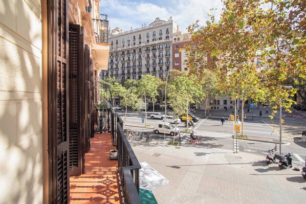 A balcony with a tiled floor, a potted plant, and a view of a city street with buildings and parked cars.