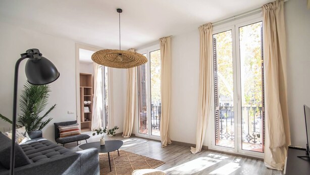 Ett rymligt vardagsrum med en modern soffa, ett runt soffbord och en hängande, vävd lampa. Stora fönster med gardiner släpper in naturligt ljus i rummet.
