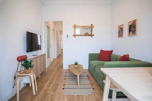 A living room with a green sofa, a wooden table, a TV mounted on the wall, and a small plant on a side table.