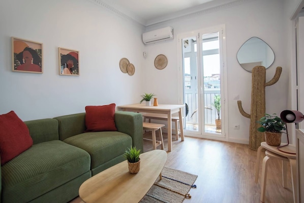 A living room with a green sofa, a wooden coffee table, and a dining area with a small table and chairs. There is a large mirror on the wall and a potted plant on the coffee table.