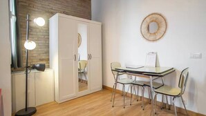 A room with a wooden floor, a white wardrobe, a round mirror, a table with four chairs, and a floor lamp.
