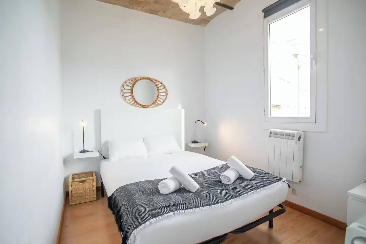 A modern bedroom with a large bed, a bedside table with a lamp, a round mirror, and a window providing natural light.