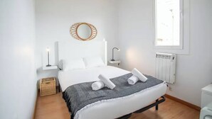 A modern bedroom with a large bed, a bedside table with a lamp, a round mirror, and a window providing natural light.