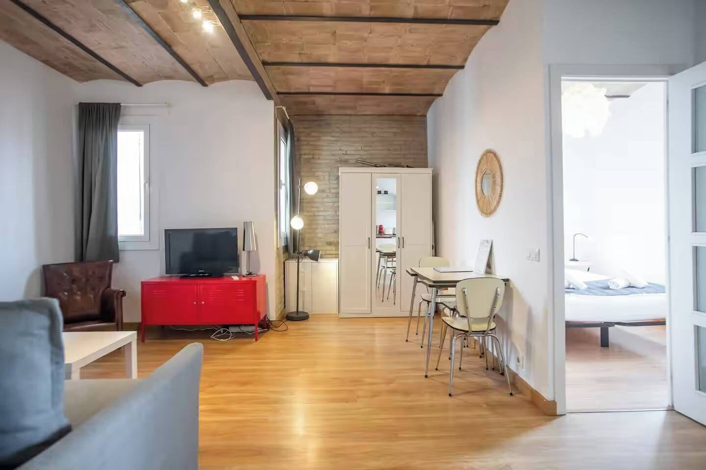 A modern living room with a wooden ceiling, a red entertainment unit, a television, a dining area with a table and chairs, and a view of a bedroom through an open door.