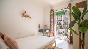 A bedroom with a bed, a desk, a chair, a potted plant, and a balcony with a view of buildings. - 12par1003- Luminoso piso en Paralelo (Barcelona)