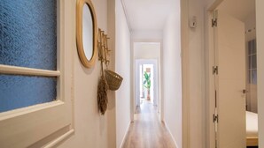 A hallway with a blue textured wall, a wooden oval mirror, a woven basket, and a wooden floor leading to a room with a bed. - 12par1003- Luminoso piso en Paralelo (Barcelona)