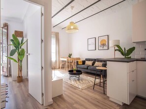 Un salon moderne avec un canapé, une table basse et un luminaire suspendu. On y trouve également un coin repas avec une table et des chaises, ainsi qu’une cuisine équipée de placards et d’un plan de travail.