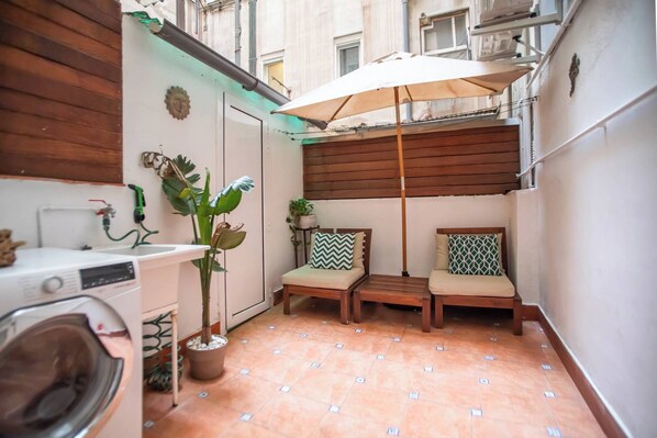 Een compact stadappartement met een klein balkon, een wasmachine, een potplant en twee fauteuils met kussens.