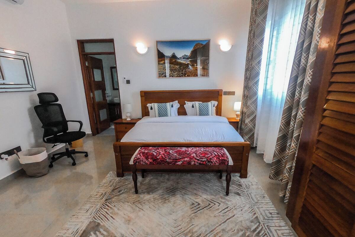 A bedroom with a bed, a chair, a rug, a painting, and a window with curtains.