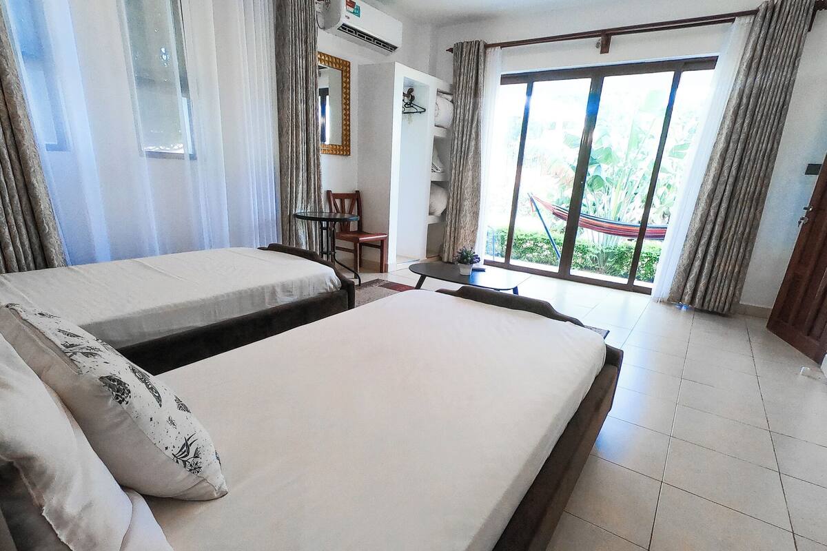 A hotel room with two beds, a small table, a chair, a window with curtains, and a view of greenery outside.