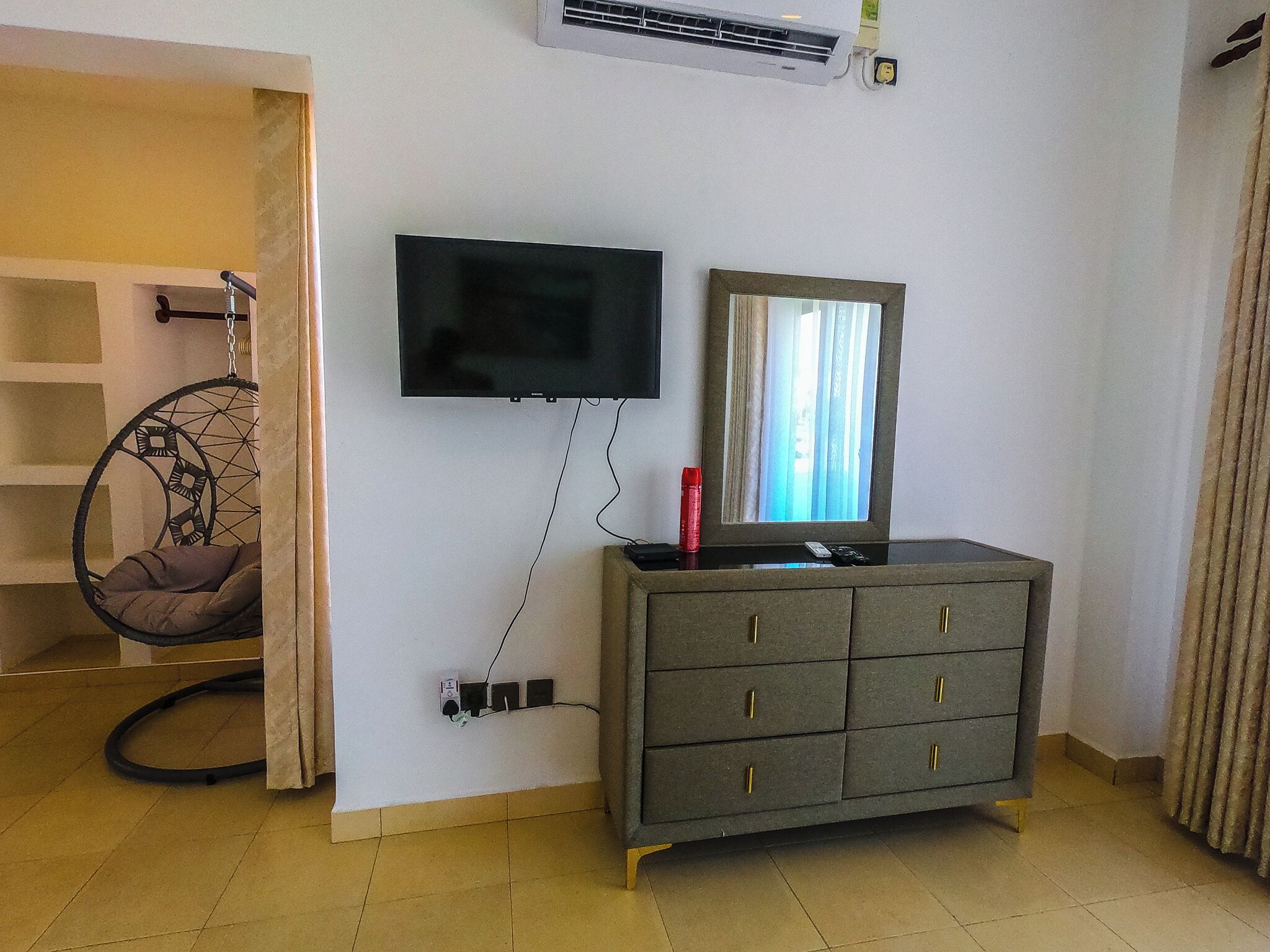 Ein Zimmer mit an der Wand befestigtem Fernseher, einem Schrank mit Schubladen, einem Hängesessel und einem Fenster mit Vorhängen.
